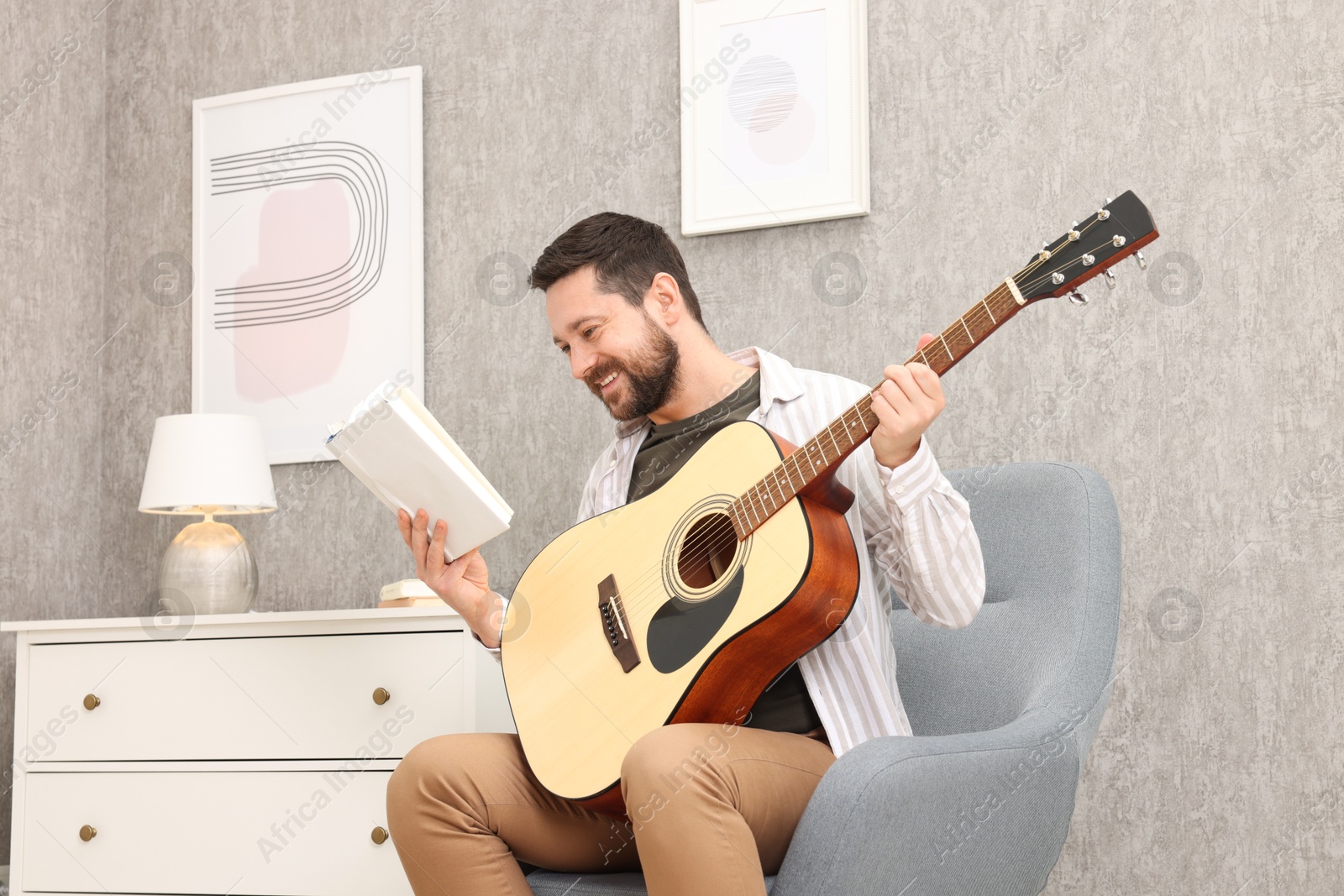 Photo of Relaxing hobby. Smiling man with guitar and book on armchair at home