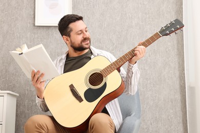 Photo of Relaxing hobby. Man with guitar and book on armchair at home