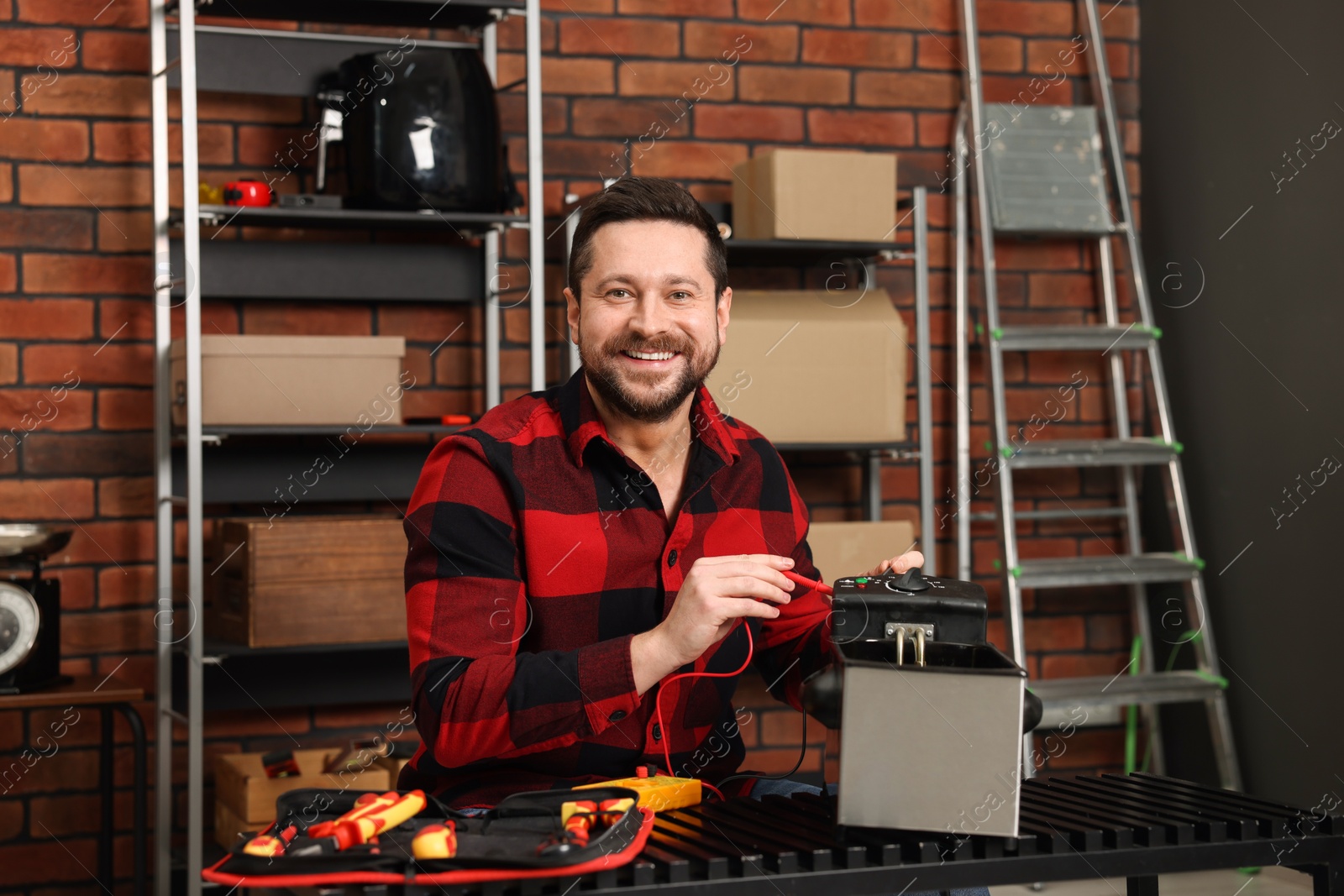 Photo of Relaxing hobby. Smiling man testing deep fryer with multimeter in workshop