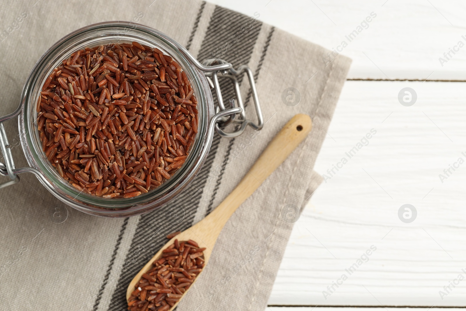 Photo of Raw brown rice in open jar and spoon on white wooden table, top view. Space for text