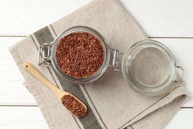 Photo of Raw brown rice in open jar and spoon on white wooden table, top view