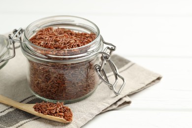 Photo of Raw brown rice in open jar and spoon on white wooden table, closeup. Space for text