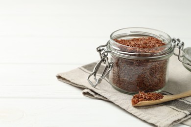 Photo of Raw brown rice in open jar and spoon on white wooden table, closeup. Space for text