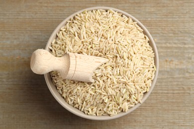 Photo of Raw brown rice and scoop in bowl on wooden table, top view