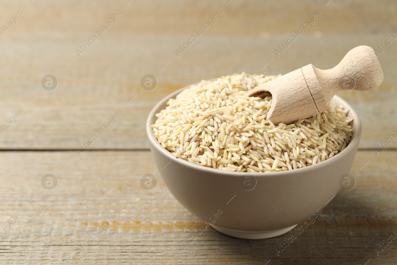 Photo of Raw brown rice and scoop in bowl on wooden table, closeup. Space for text