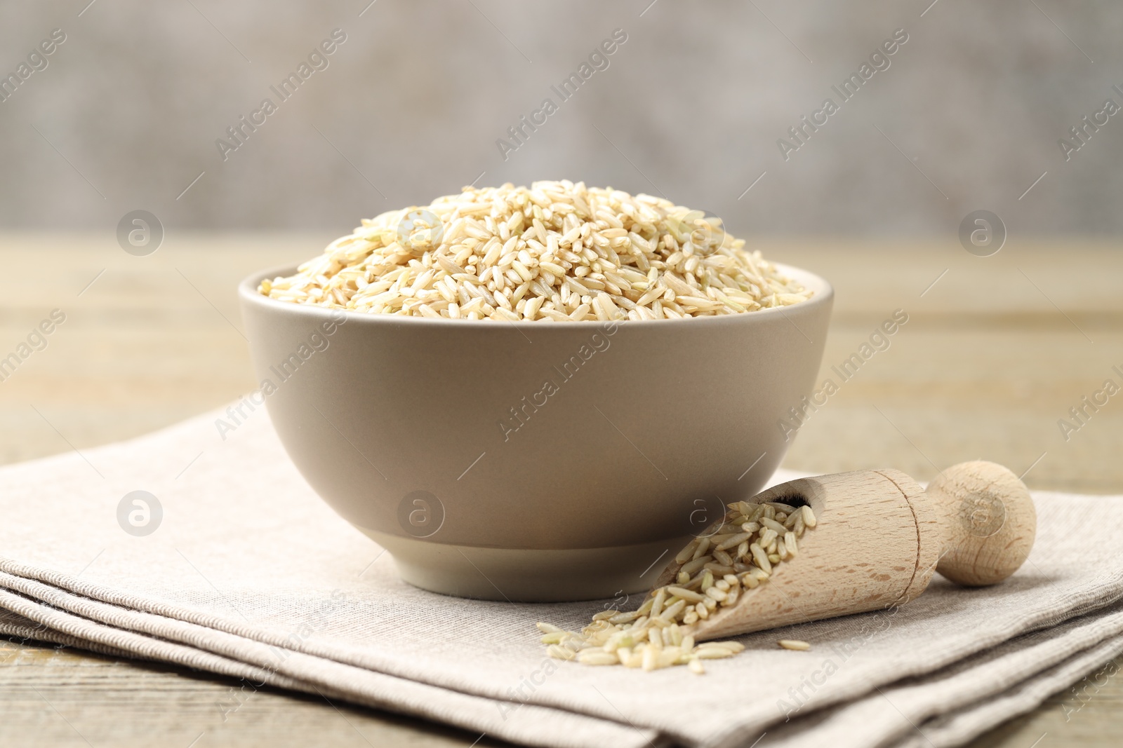 Photo of Raw brown rice in bowl and scoop on wooden table against grey background, closeup