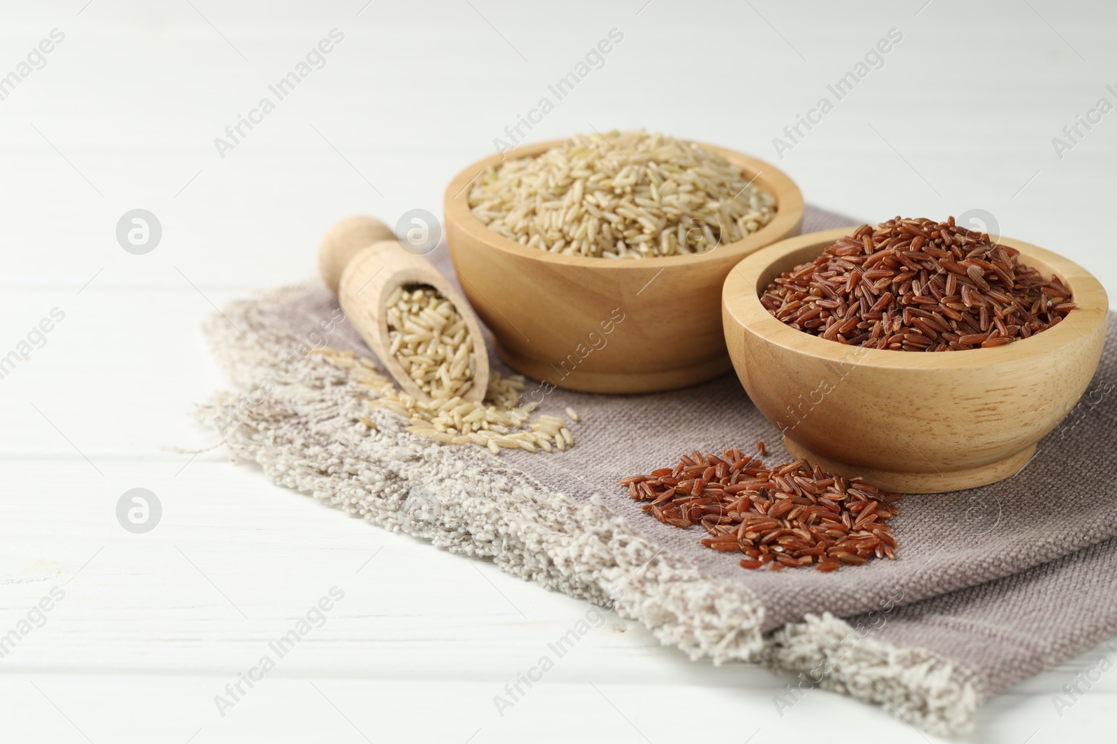 Photo of Different types of brown rice in bowls and scoop on white wooden table, closeup. Space for text