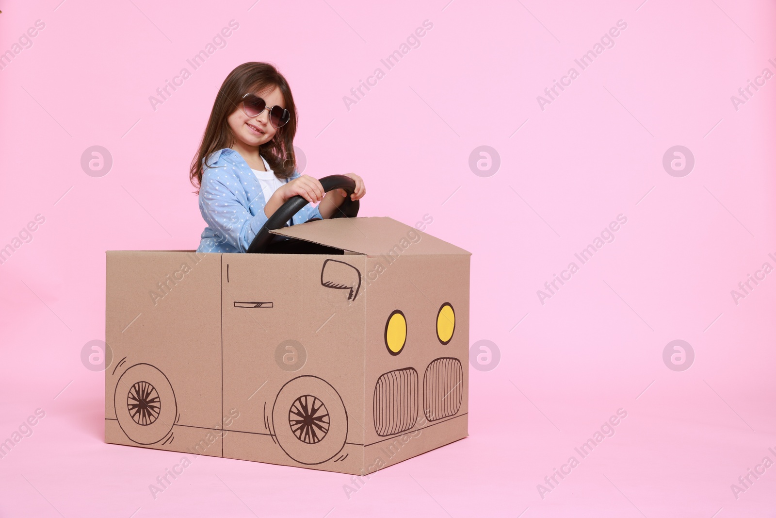 Photo of Little girl in sunglasses driving car made with cardboard on light pink background. Space for text