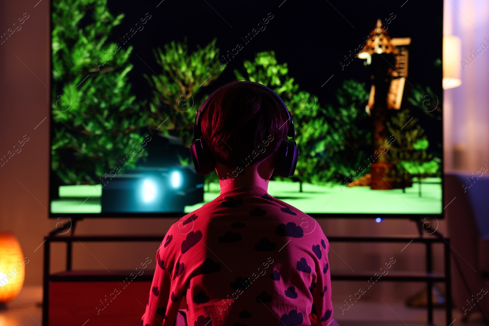 Photo of Little boy in headphones playing video game indoors at night, back view