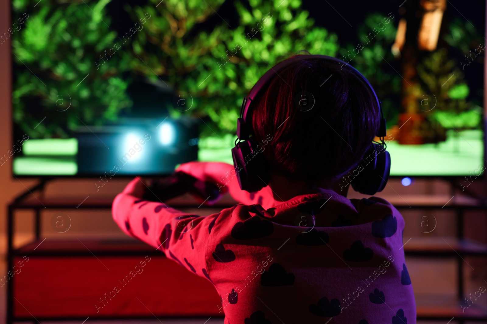 Photo of Little boy in headphones playing video game indoors at night, back view