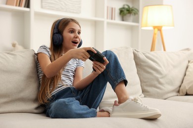 Photo of Cute girl in headphones playing video game on sofa at home