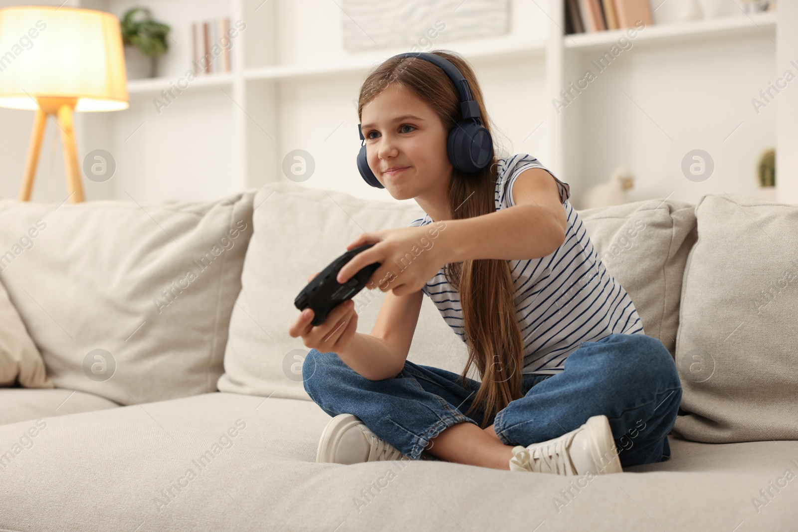 Photo of Cute girl in headphones playing video game on sofa at home