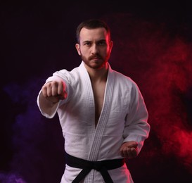 Photo of Man wearing uniform practicing karate in color lights and smoke against black background