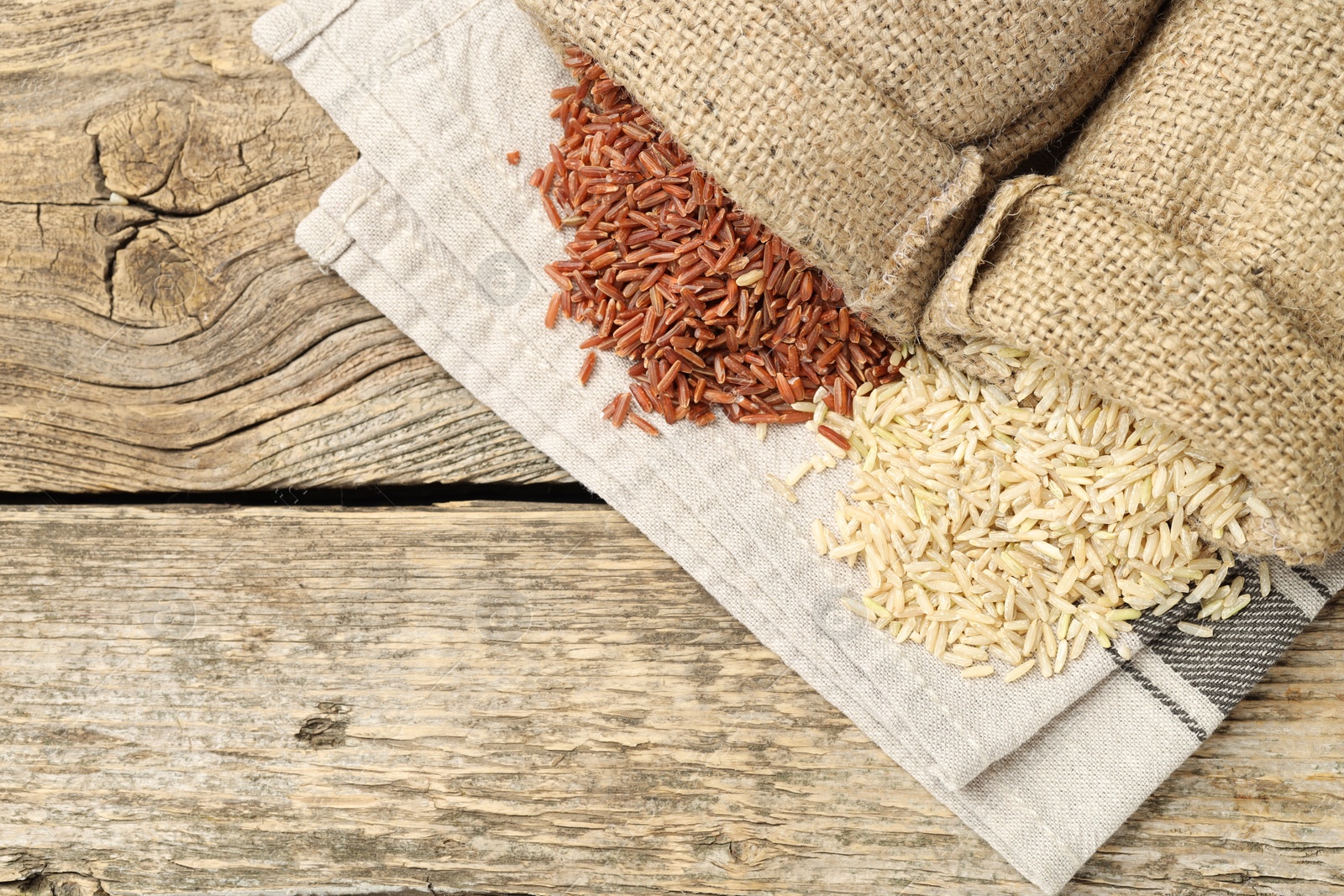 Photo of Different types of brown rice in sacks on wooden table, flat lay. Space for text