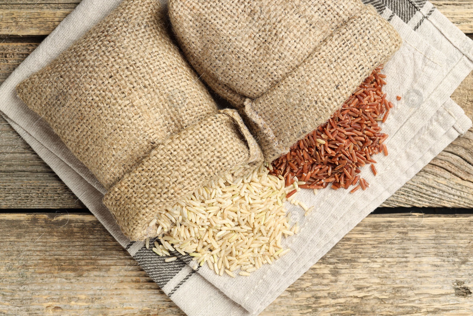 Photo of Different types of brown rice in sacks on wooden table, flat lay