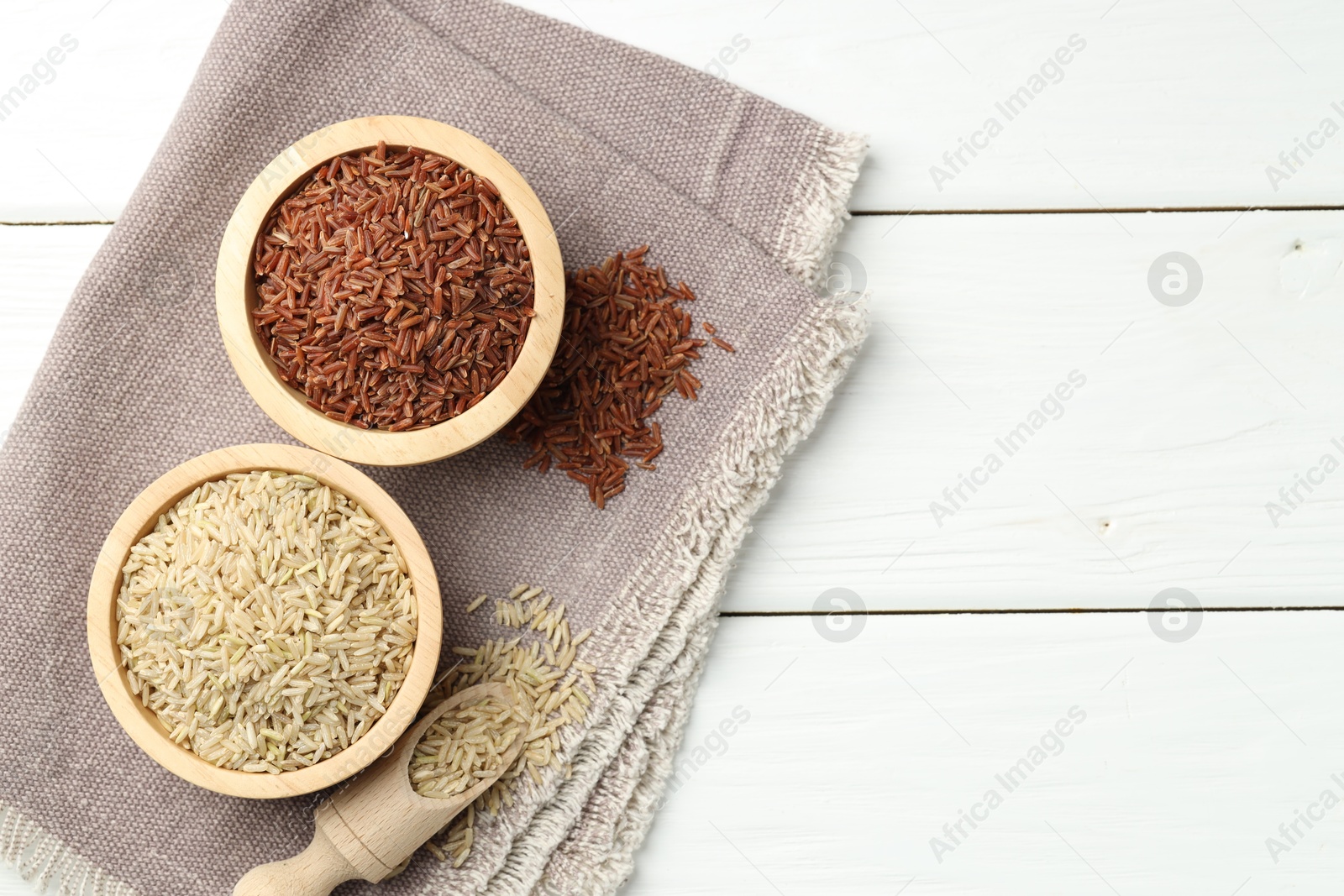 Photo of Different types of brown rice in bowls and scoop on white wooden table, flat lay. Space for text