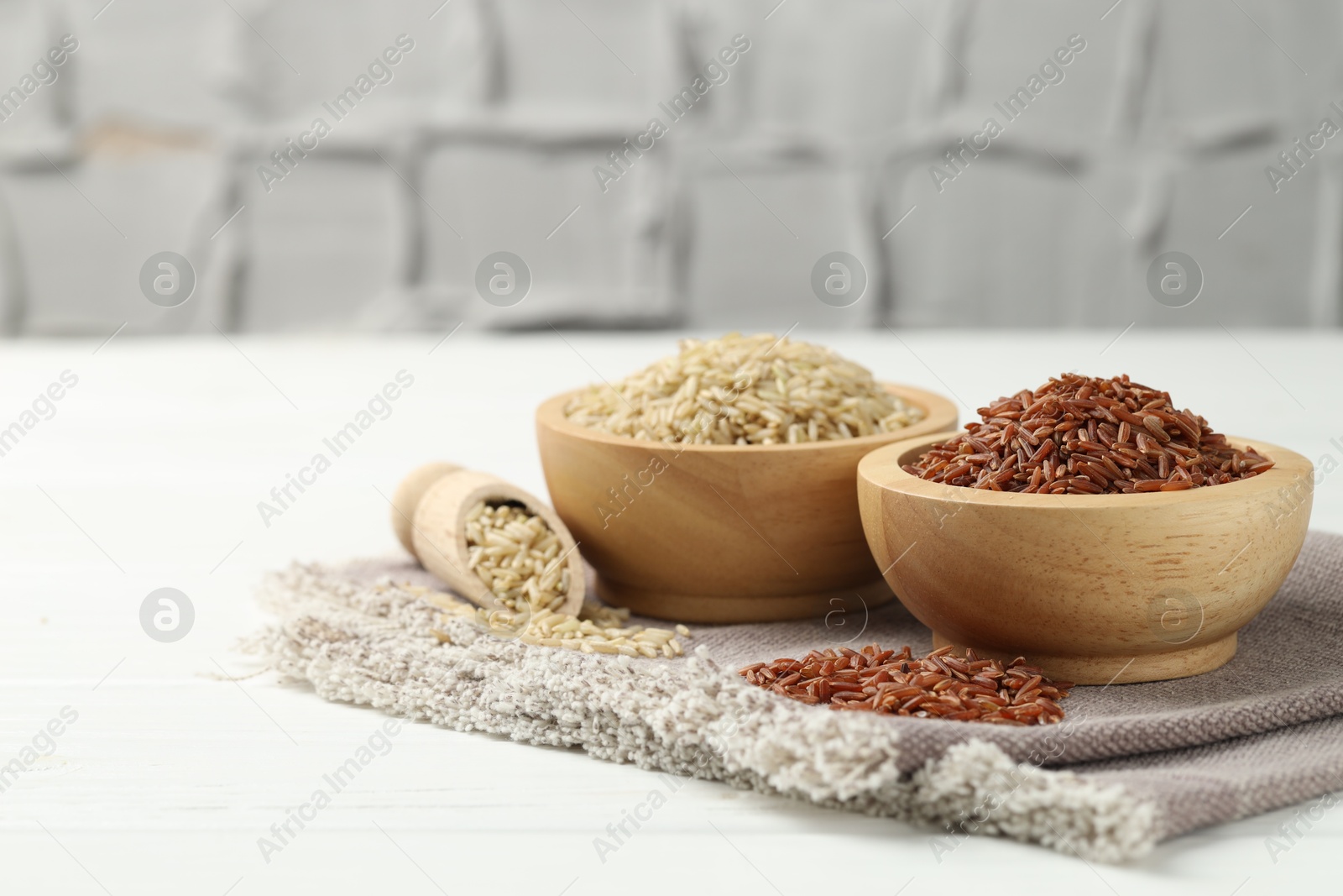 Photo of Different types of brown rice in bowls and scoop on white wooden table, closeup. Space for text