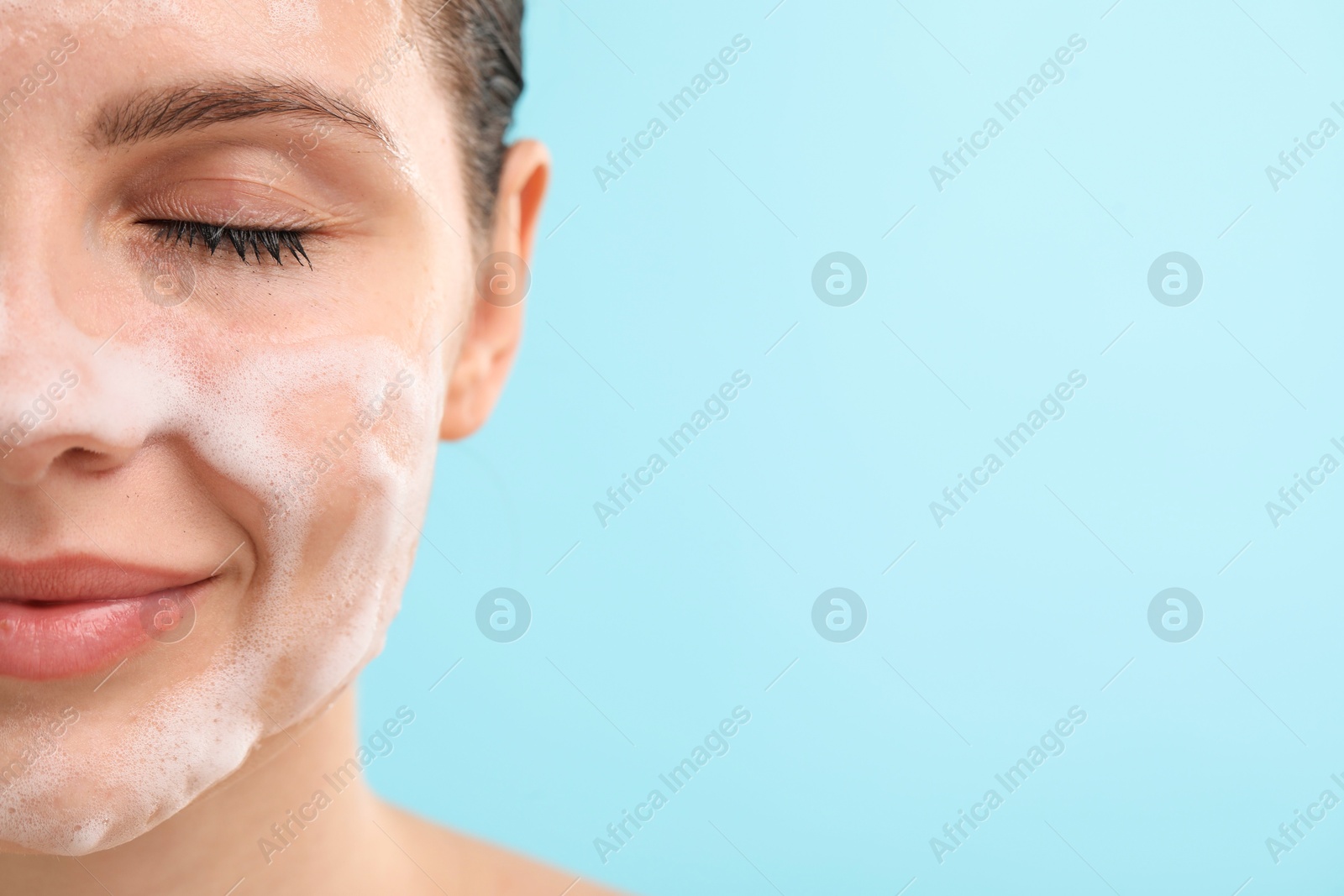 Photo of Attractive woman washing her face with cleansing foam on light blue background, closeup. Space for text