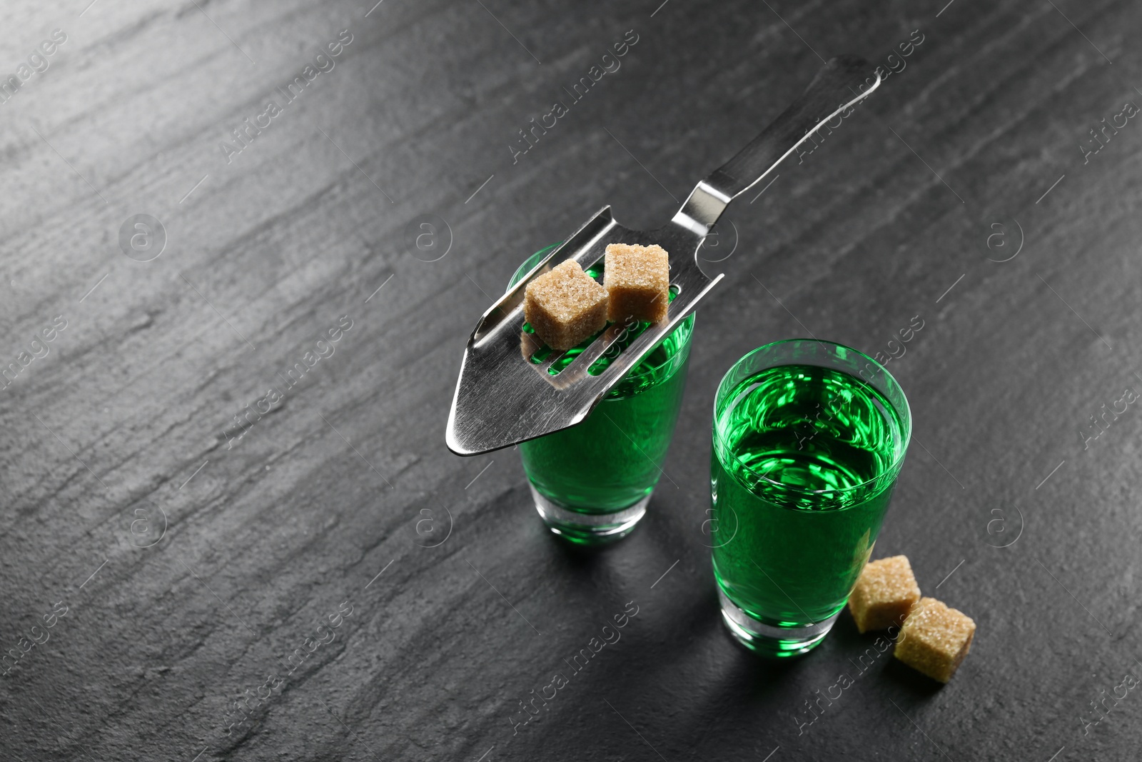 Photo of Absinthe in shot glasses, brown sugar and slotted spoon on black table, closeup. Space for text