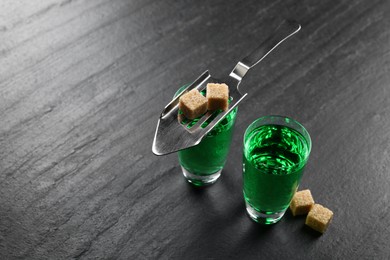 Photo of Absinthe in shot glasses, brown sugar and slotted spoon on black table, closeup. Space for text