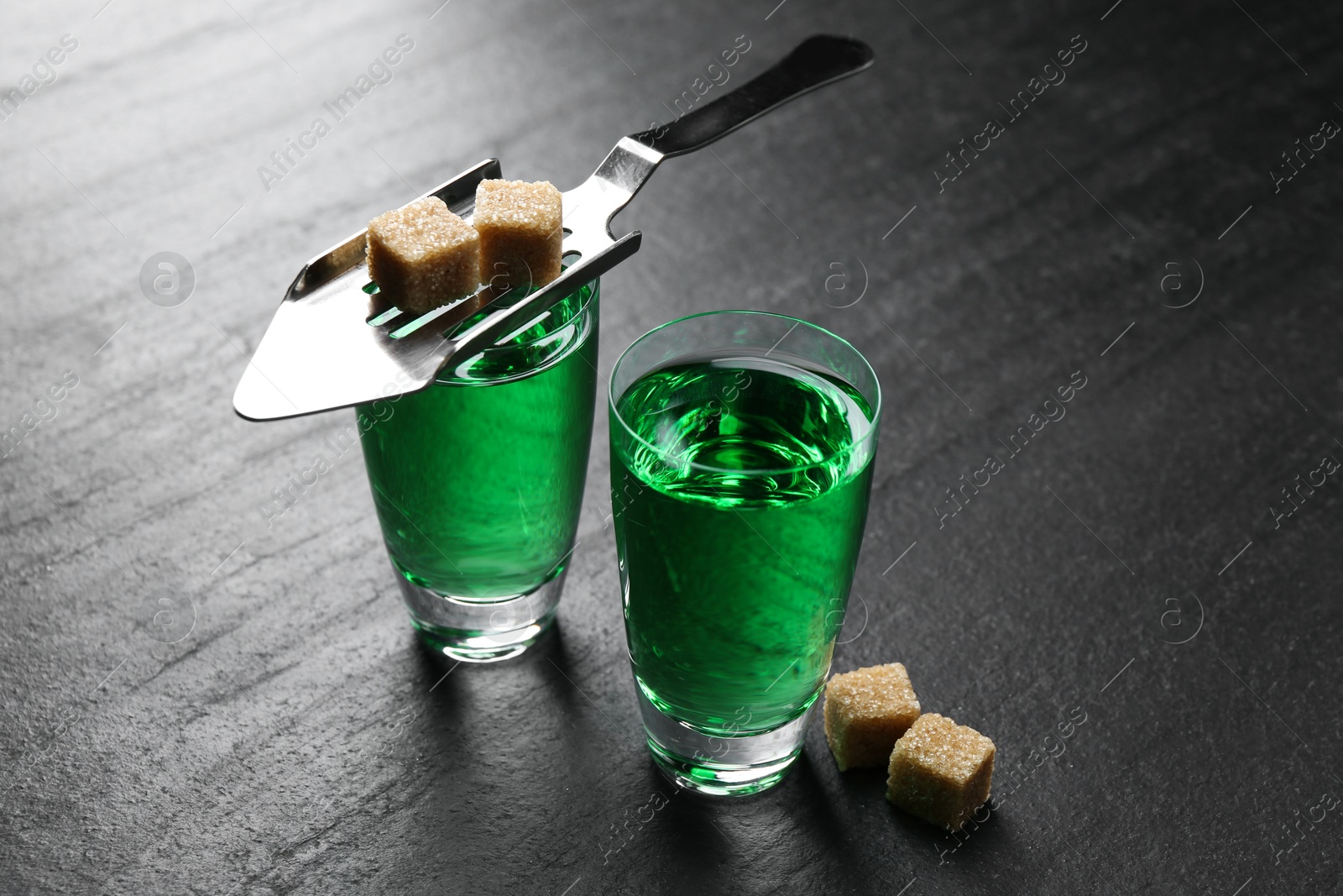 Photo of Absinthe in shot glasses, brown sugar and slotted spoon on black table, closeup