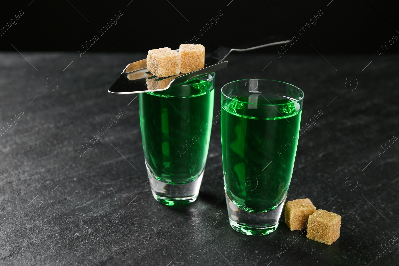 Photo of Absinthe in shot glasses, brown sugar and slotted spoon on black table, closeup