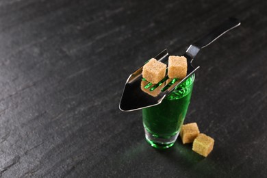 Photo of Absinthe in shot glass, brown sugar and slotted spoon on black table, closeup. Space for text