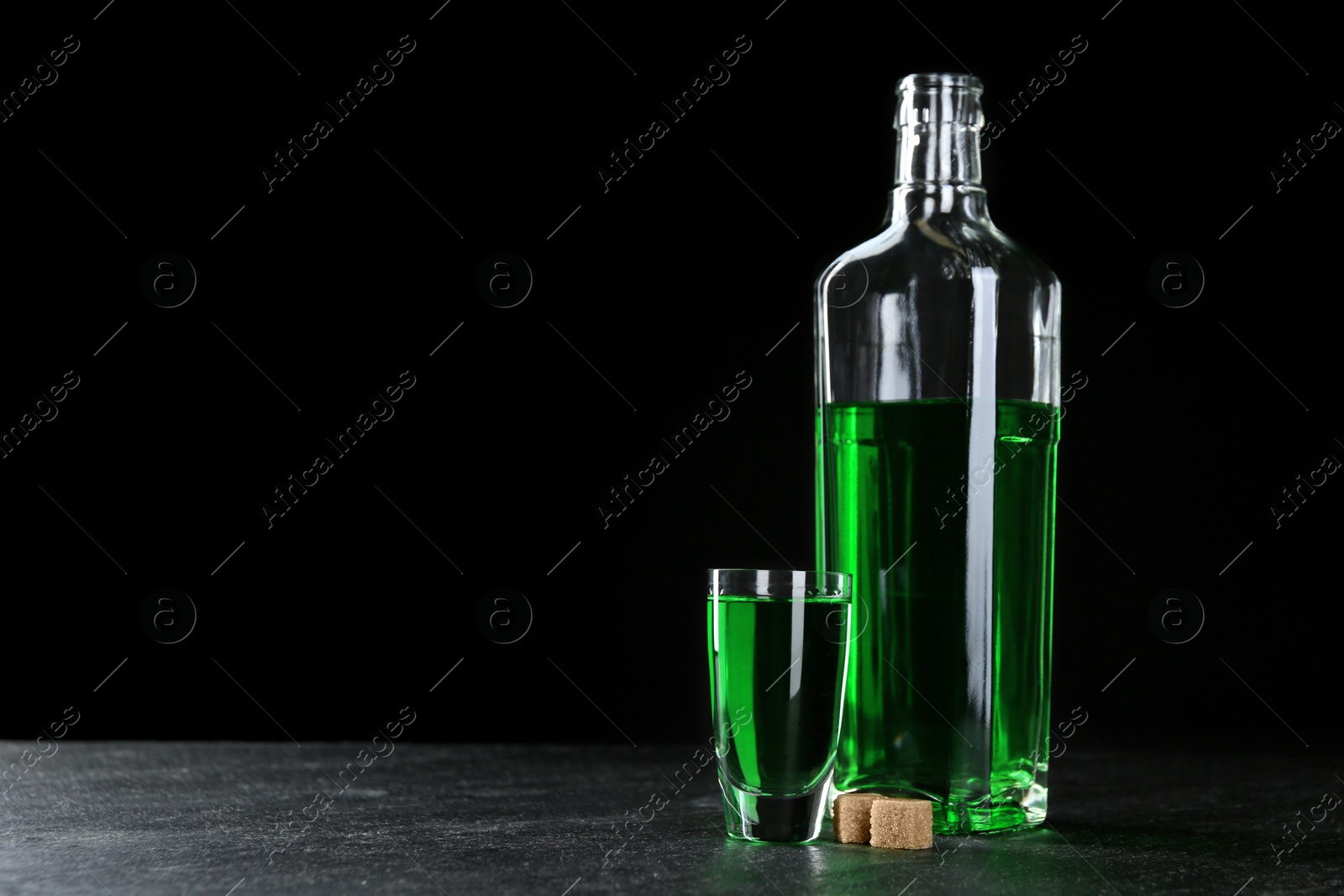 Photo of Absinthe in shot glass, bottle and brown sugar on table against black background, space for text