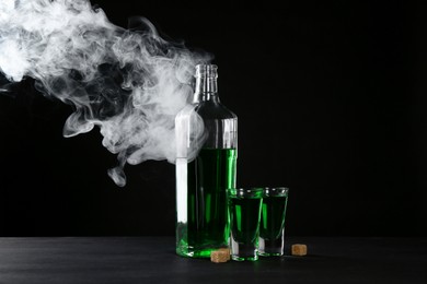 Photo of Absinthe in shot glasses, bottle and brown sugar on table against black background with smoke