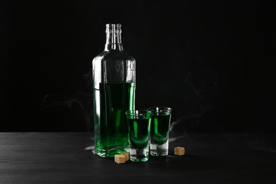 Photo of Absinthe in shot glasses, bottle and brown sugar on table against black background with smoke