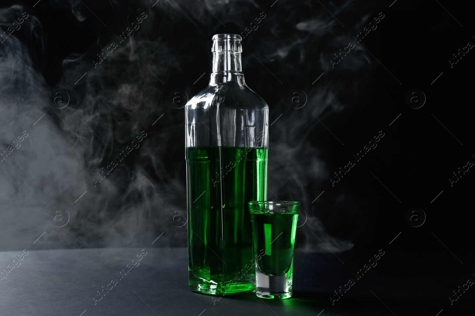 Photo of Absinthe in shot glass and bottle on table against black background with smoke