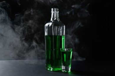 Photo of Absinthe in shot glass and bottle on table against black background with smoke