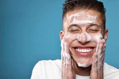 Photo of Smiling man washing his face with cleansing foam on blue background, closeup. Space for text