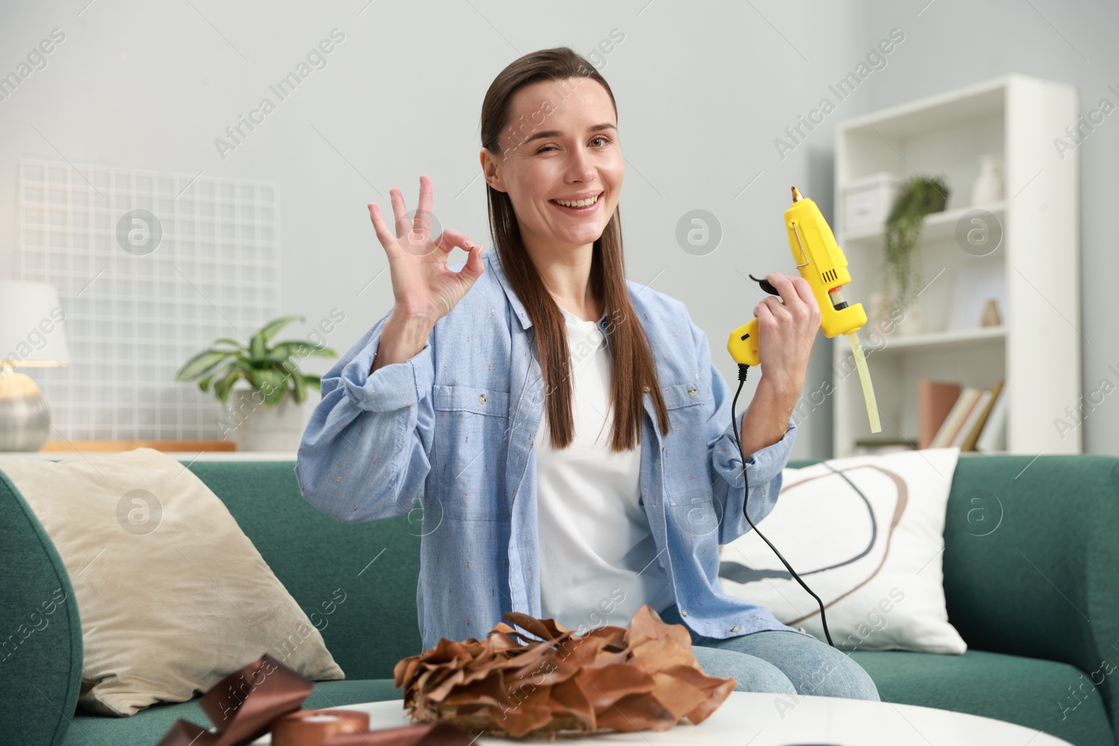 Photo of Woman with hot glue gun showing OK gesture at table indoors