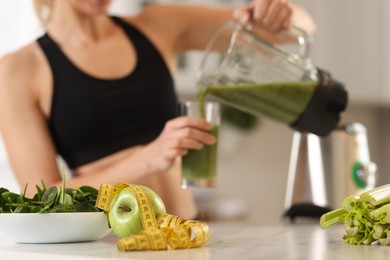 Photo of Weight loss. Woman pouring fresh shake from blender cup into glass, focus on ingredients and measuring tape