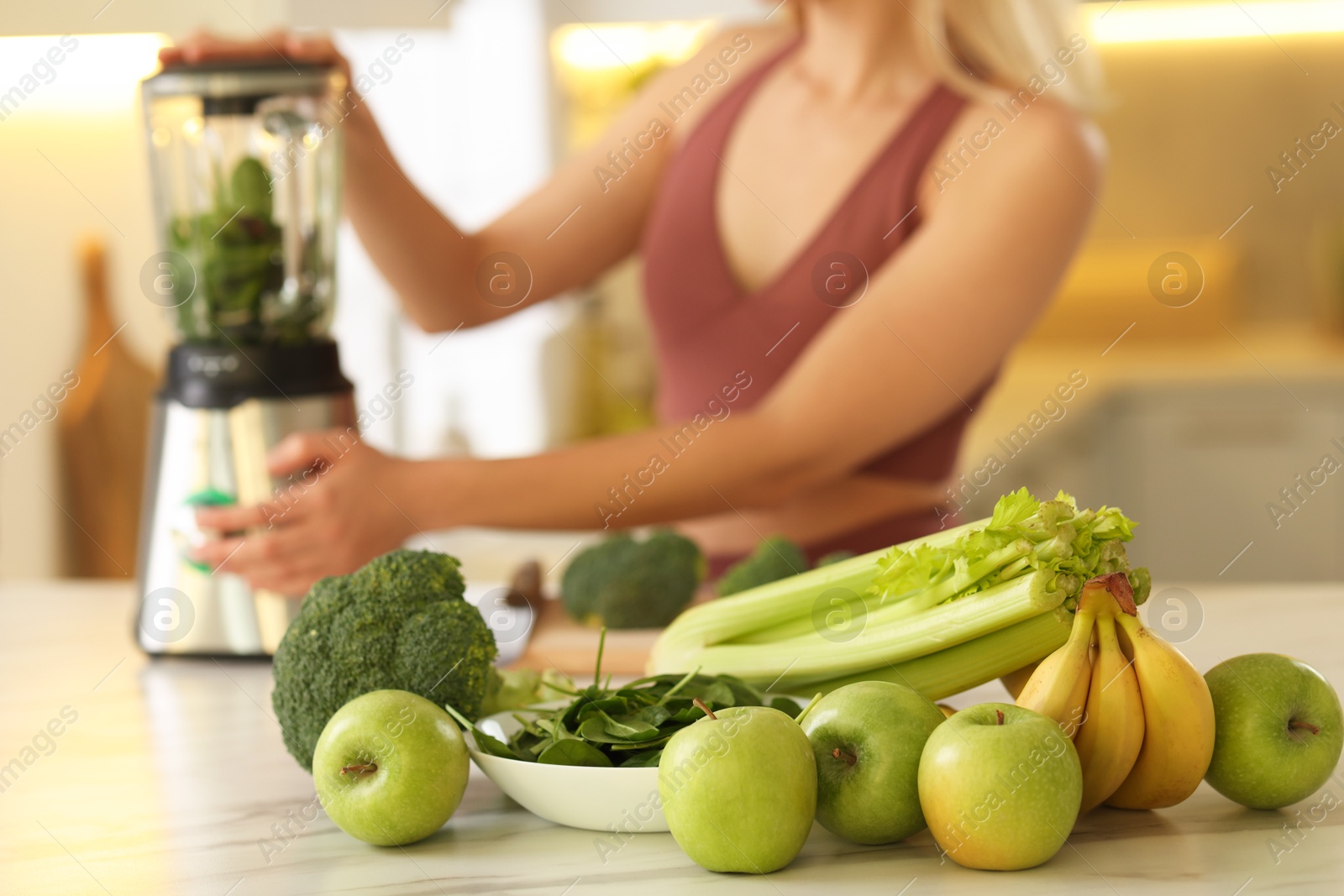 Photo of Weight loss. Woman making protein shake at table in kitchen, focus on fresh vegetables and fruits