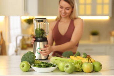 Photo of Weight loss. Woman making protein shake at table in kitchen, focus on fresh vegetables and fruits