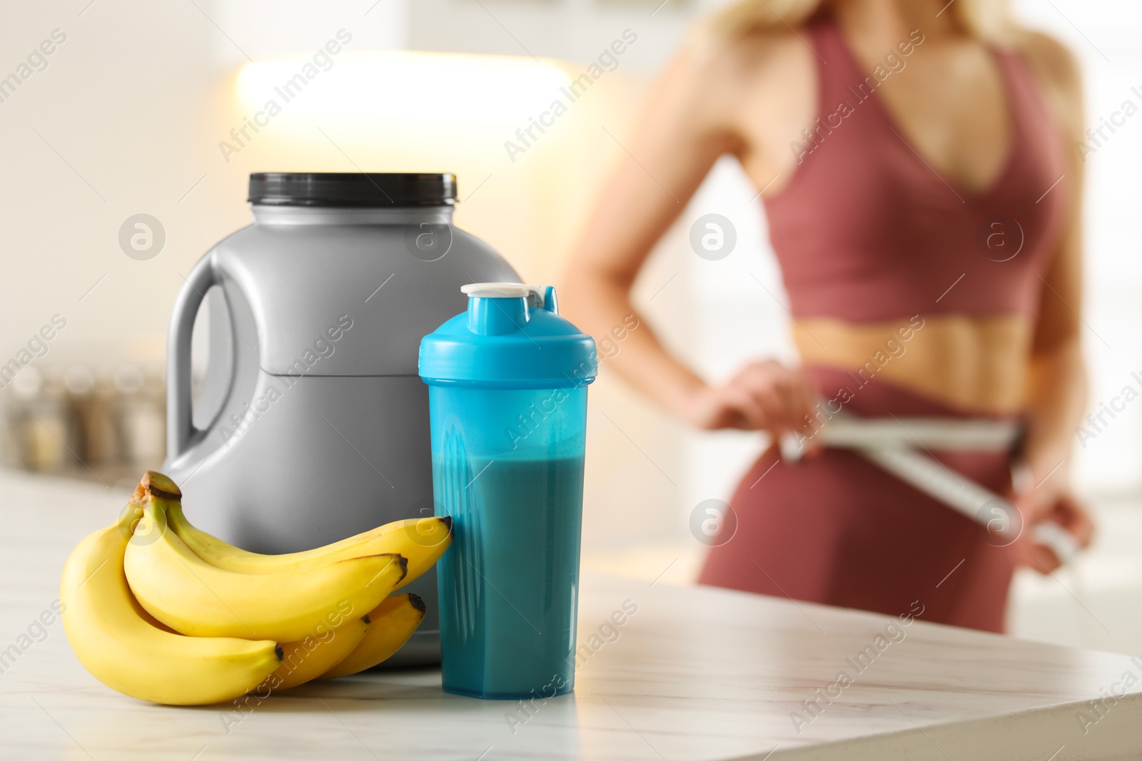 Photo of Weight loss. Woman measuring waist with tape in kitchen, focus on bananas, shaker of protein and jar