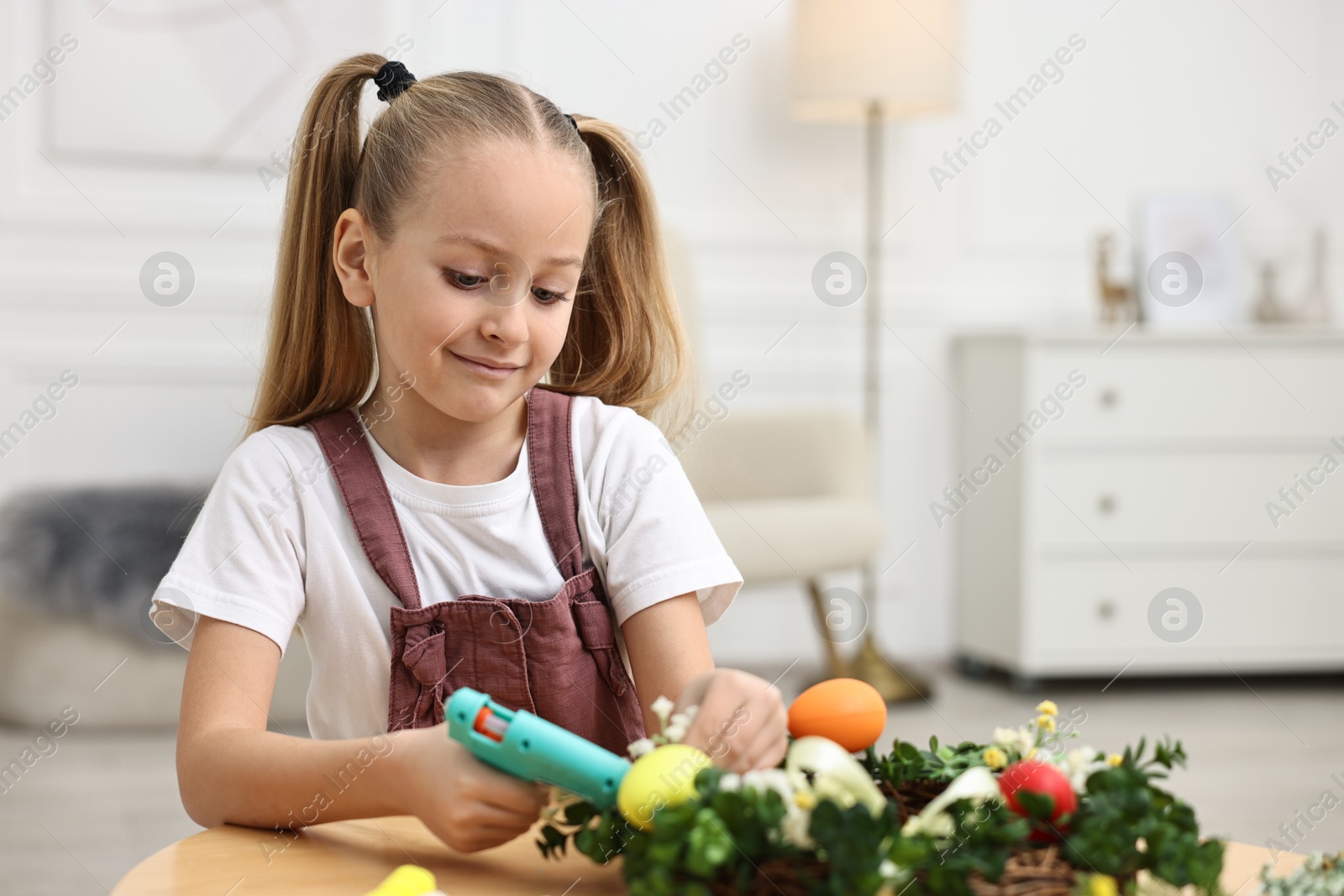 Photo of Little girl with hot glue gun creating Easter composition at table indoors