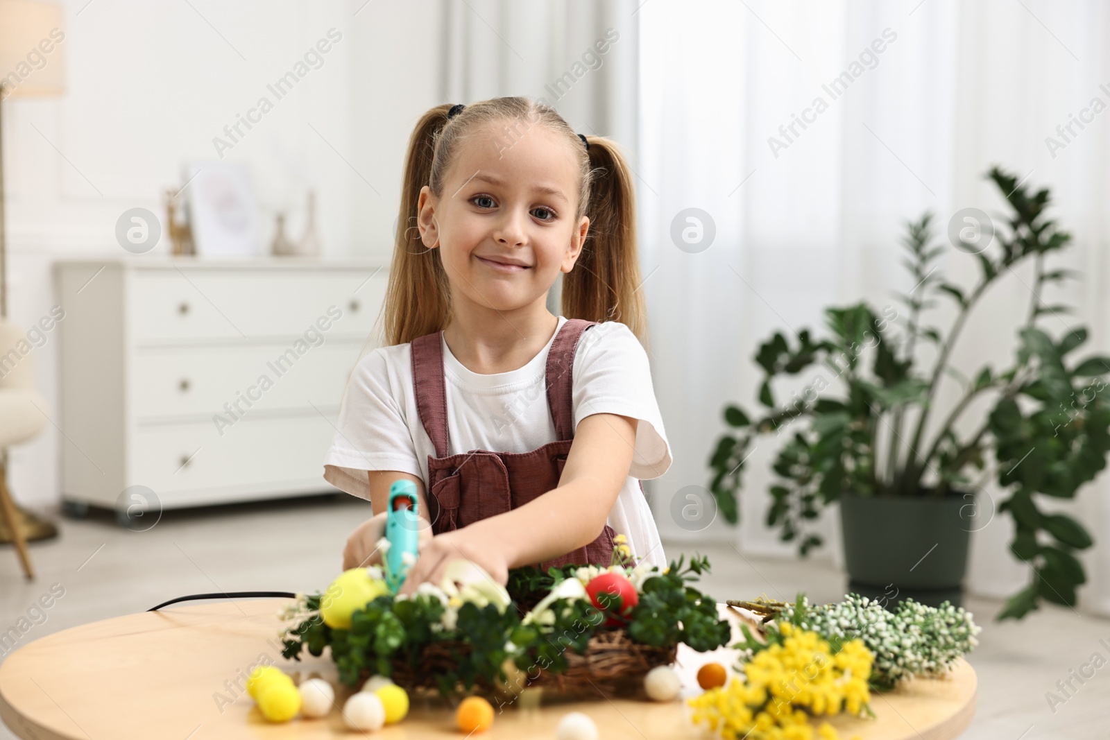 Photo of Little girl with hot glue gun creating Easter composition at table indoors