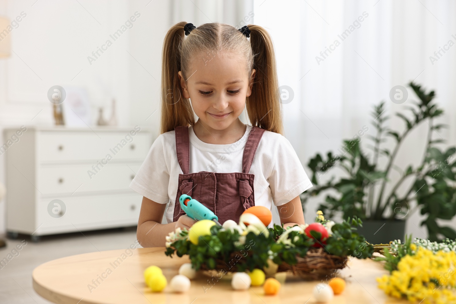 Photo of Little girl with hot glue gun creating Easter composition at table indoors