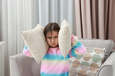 Photo of Angry little girl covering her ears with pillows from loud sound on armchair at home