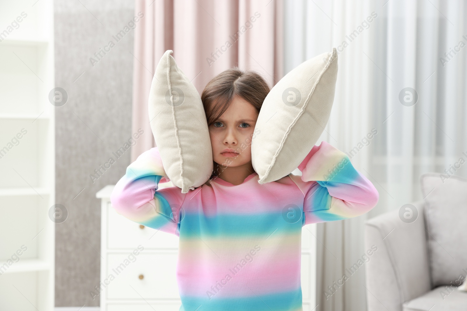 Photo of Exhausted little girl covering her ears with pillows from loud sound at home