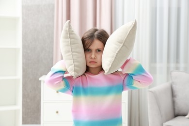 Photo of Exhausted little girl covering her ears with pillows from loud sound at home