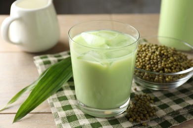 Photo of Fresh mung bean juice with ice in glass, leaves and seeds on wooden table, closeup