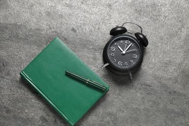 Photo of Time for knowledge. Alarm clock, notebook and pen on grey textured table, flat lay