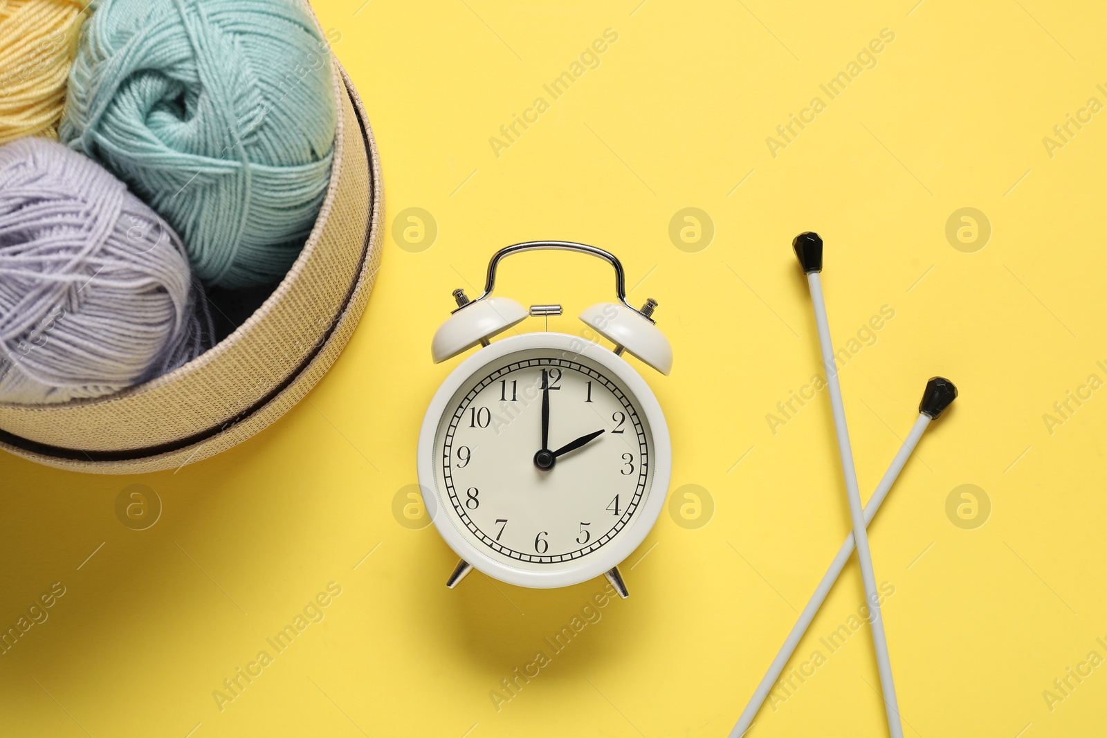 Photo of Time for hobby. Alarm clock, yarn and knitting needles on yellow background, flat lay