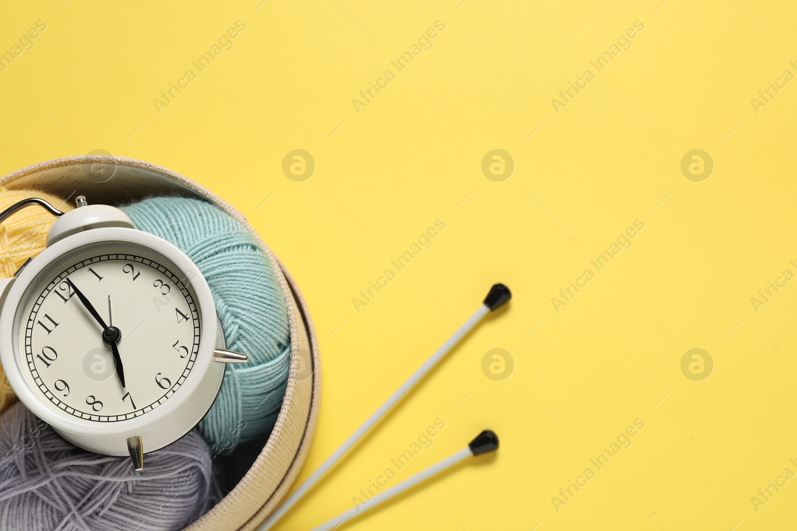 Photo of Time for hobby. Alarm clock, yarn and knitting needles on yellow background, flat lay. Space for text