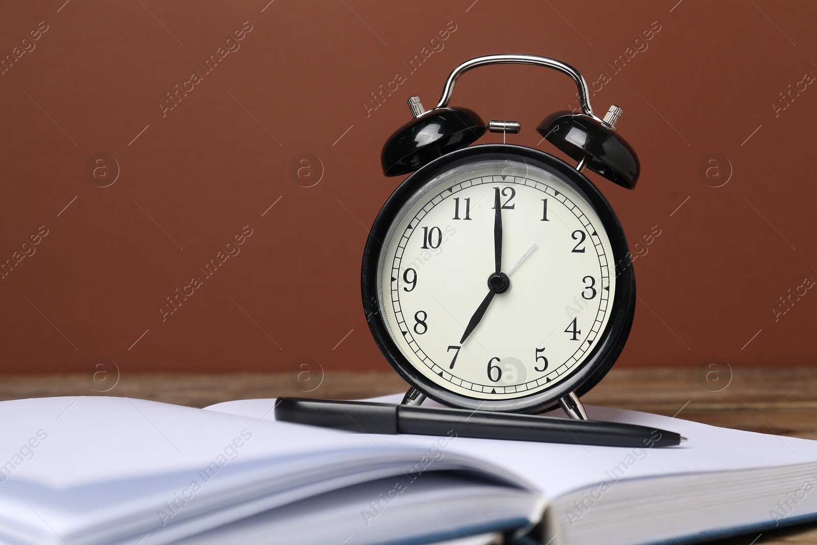 Photo of Time for knowledge. Alarm clock, book and pen on wooden table, closeup