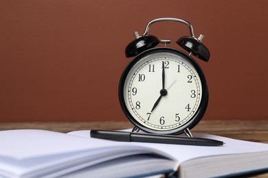 Photo of Time for knowledge. Alarm clock, book and pen on wooden table, closeup